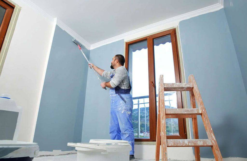 A man in overalls uses a roller to paint a light blue wall near a window in a room under renovation.