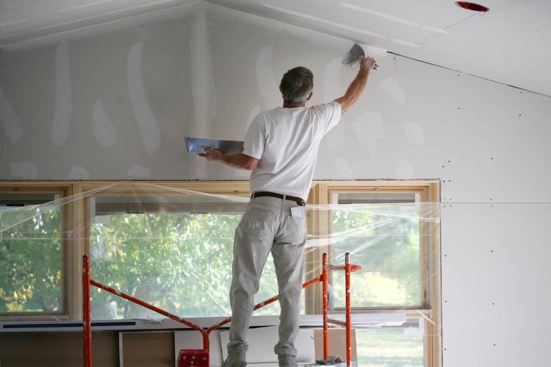 A man stands on a ladder applying joint compound to a drywall ceiling with a putty knife.