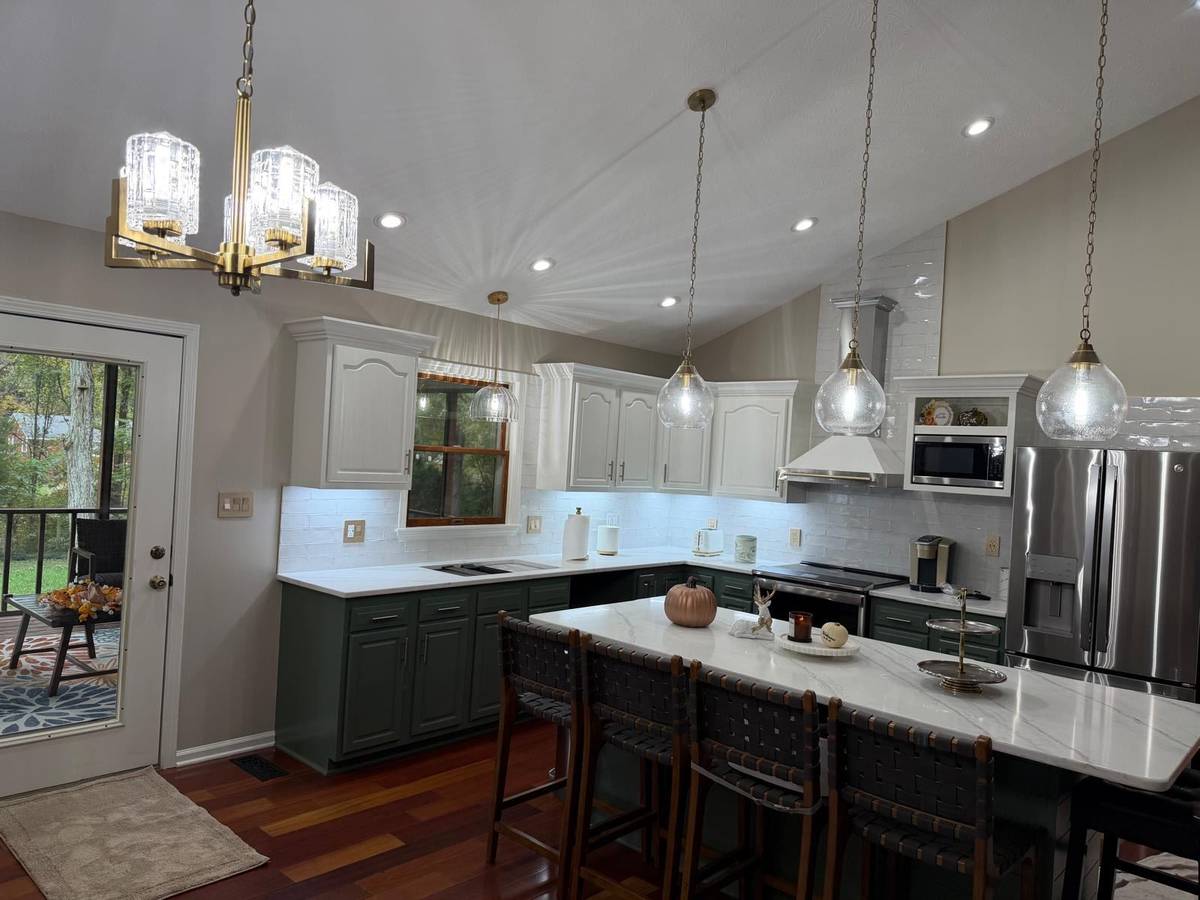 A contemporary kitchen featuring white cabinets, dark green lower cabinets, a marble island with woven bar stools, and pendant lighting.